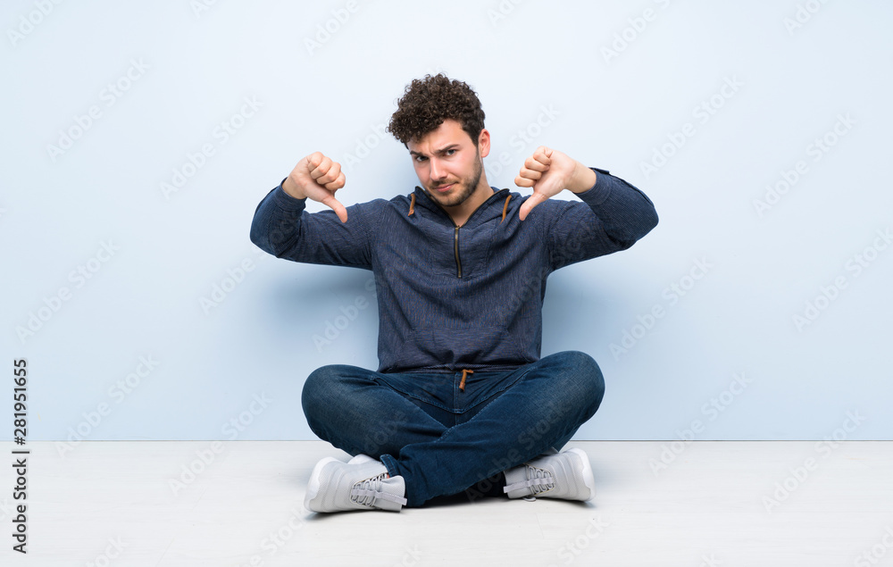 Young man sitting on the floor showing thumb down Young man sitting on the floor showing thumb down