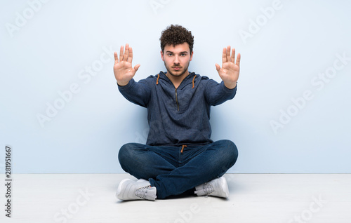 Young man sitting on the floor making stop gesture and disappointed