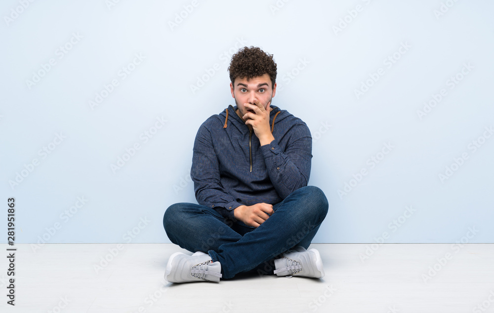 Young man sitting on the floor surprised and shocked while looking right Young man sitting on the floor surprised and shocked while looking right