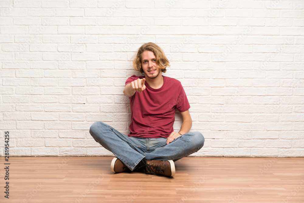 Blonde man sitting on the floor points finger at you with a confident expression Blonde man sitting on the floor points finger at you with a confident expression
