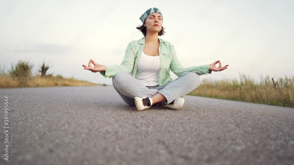 Young trendy woman meditating in lotus pose sitting on empty road between desert sands. Yoga, religion, zen concept. Young trendy woman meditating in lotus pose sitting on empty road between desert sands. Yoga, religion, zen concept.