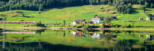 Train passing a traditional Norwegian summer farm along lake Vangsvatnet reflected in the water near Voss Hordaland county Norway © HildaWeges