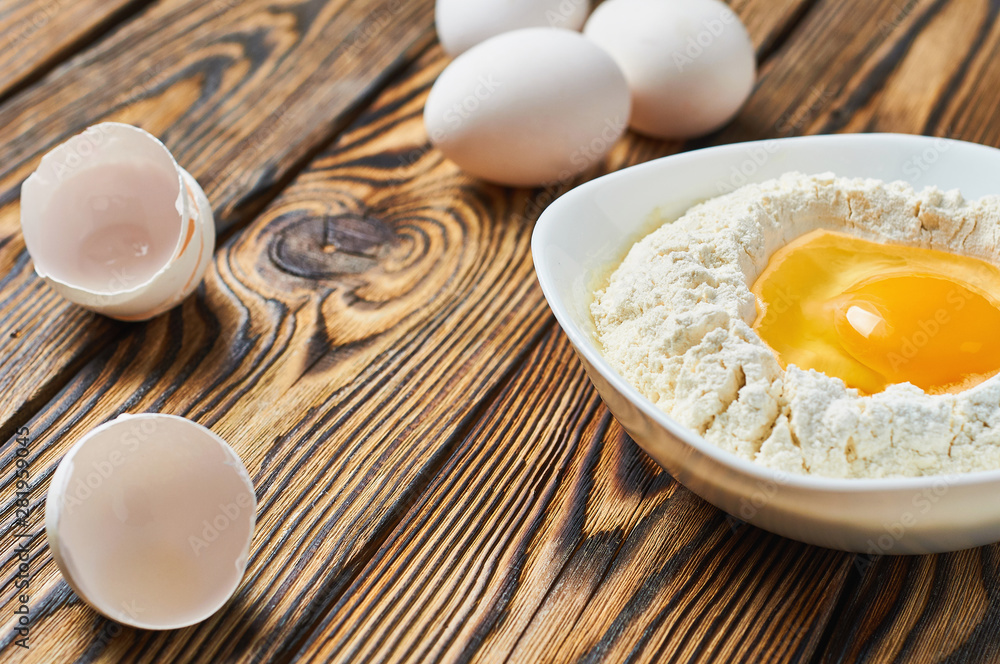Broken egg in ceramic bowl with flour lies near shells on rustic wooden table. Space for text Broken egg in ceramic bowl with flour lies near shells on rustic wooden table. Space for text
