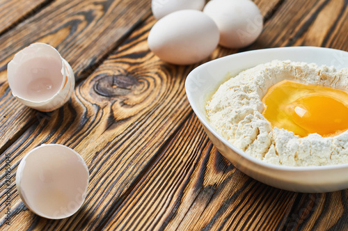 Broken egg in ceramic bowl with flour lies near shells on rustic wooden table. Space for text