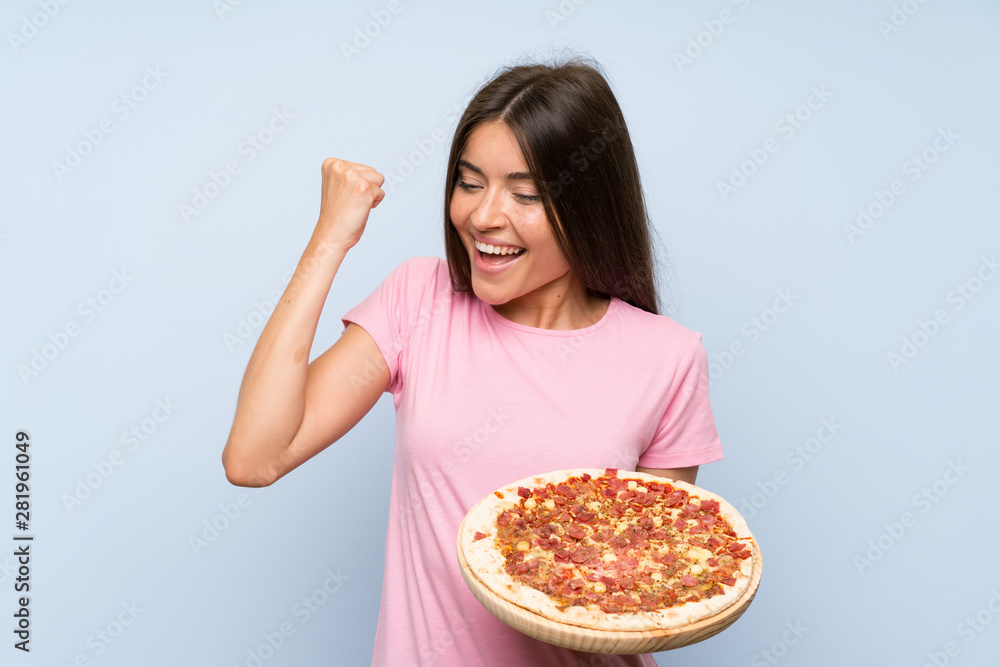 Pretty young girl holding a pizza over isolated blue wall celebrating a victory