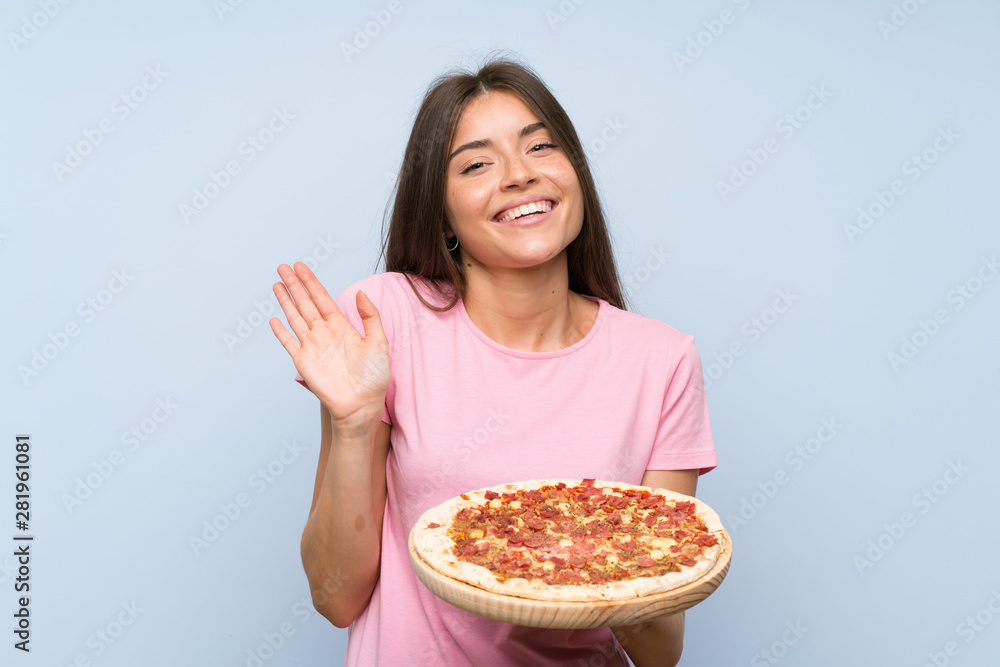 Pretty young girl holding a pizza over isolated blue wall saluting with hand with happy expression