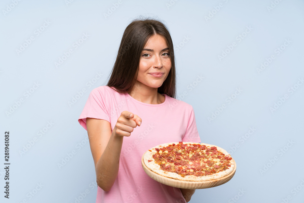 Pretty young girl holding a pizza over isolated blue wall points finger at you with a confident expression