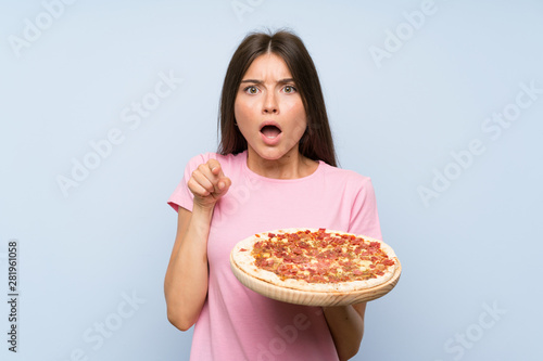 Pretty young girl holding a pizza over isolated blue wall surprised and pointing front