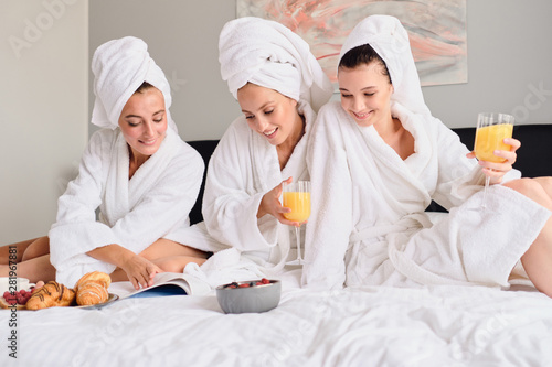 Young beautiful women in white bathrobes and towels on head dreamily reading book together. Pretty girls holding glasses with orange juice in hands having breakfast in bed in hotel