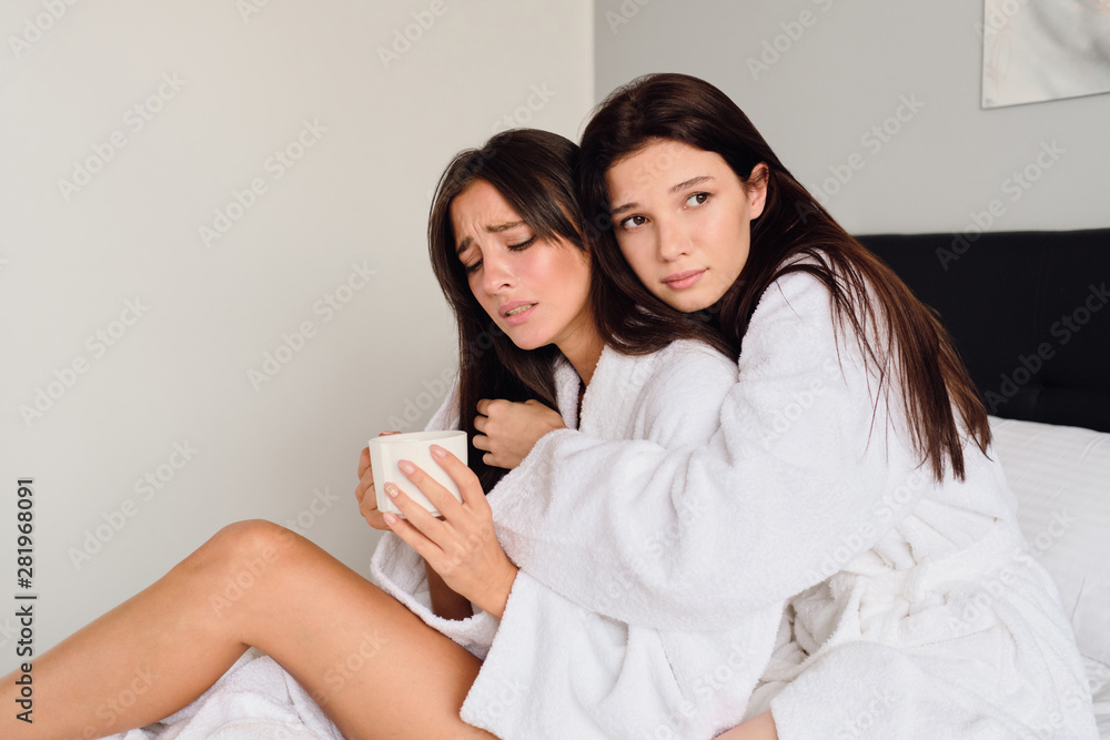 Young upset woman in white bathrobe holding cup of coffee in hand and crying while pensive girl thoughtfully hugging and supporting her on big bed at cozy home Young upset woman in white bathrobe holding cup of coffee in hand and crying while pensive girl thoughtfully hugging and supporting her on big bed at cozy home