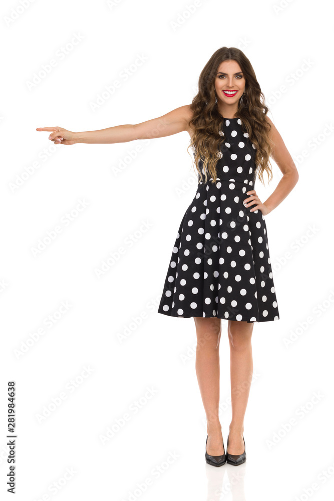 Fototapeta premium Beautiful Young Woman In Black Cocktail Dress In Polka Dots And High Heels Is Pointing And Smiling