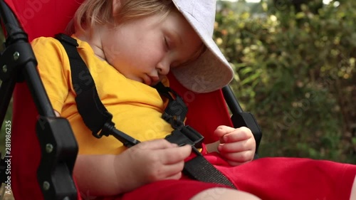 Close up outdoor portrait of little blonde boy in cap sleeping sweet in red carriage in summer beautiful forest with sun shining on background. Child health care concept. Traveling lifestyle.