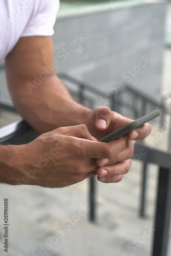  Male strong muscled hands holding black smartphone and typing message leaning on stair linen. closeup front view photography. Background of stadium stairs blurred. 