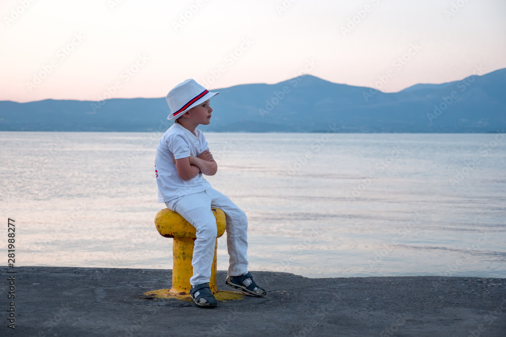 Stylish young hipster sitting on moorage waiting for ship. Little boy dressed in white clothes and hat sitting by the sea. Waiting background. Traveling concept Vacation Holiday Leisuretime Recreation Stylish young hipster sitting on moorage waiting for ship. Little boy dressed in white clothes and hat sitting by the sea. Waiting background. Traveling concept Vacation Holiday Leisuretime Recreation