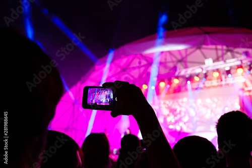 Raised hands of a man with a cell phone with a digital screen in front of bright lights record a concert. Unrecognizable people take photos and videos on the phone in front of the stage. Night show.