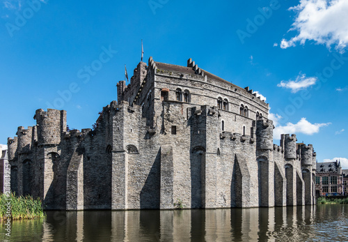 Gent, Flanders, Belgium - June 21, 2019: Gray stone castle and ramparts of Gravensteen, historic medieval castle of city, behind its moad against blue sky with white clouds. Flags on top.