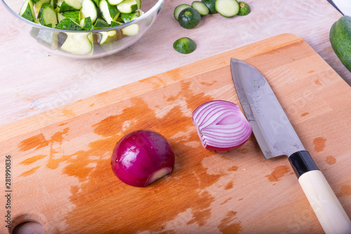 Halved red onion and santoku knife on a cutting board and chopped cucumbers in a bowl for salad. photo