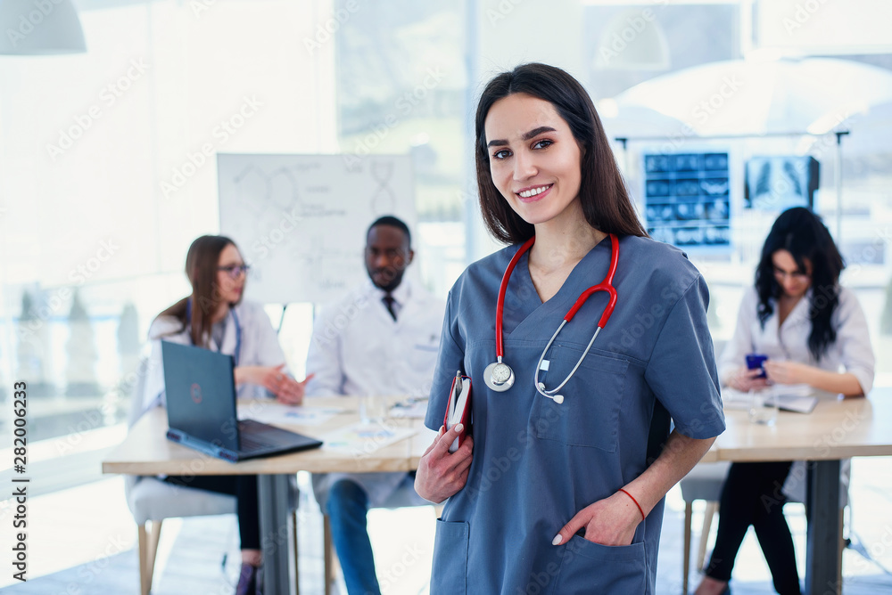 Female doctor in grey uniform with stethoscope on the neck in medical clothes smiling to the camera and crossing hands while medical team working on the background in modern hospital. Female doctor in grey uniform with stethoscope on the neck in medical clothes smiling to the camera and crossing hands while medical team working on the background in modern hospital.