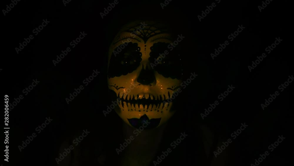 Halloween mask CALAVERA CATRINA. A girl in a blinking and frightening light. Mexican day of the dead. Portrait of a young woman with scary multi-colored makeup for Halloween on a dark background. 4K Halloween mask CALAVERA CATRINA. A girl in a blinking and frightening light. Mexican day of the dead. Portrait of a young woman with scary multi-colored makeup for Halloween on a dark background. 4K