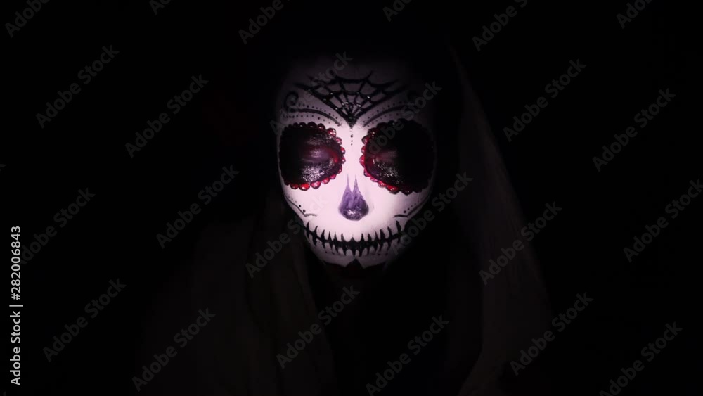 Halloween mask CALAVERA CATRINA. A girl in a blinking and frightening light. Mexican day of the dead. Portrait of a young woman with scary multi-colored makeup for Halloween on a dark background. 4K Halloween mask CALAVERA CATRINA. A girl in a blinking and frightening light. Mexican day of the dead. Portrait of a young woman with scary multi-colored makeup for Halloween on a dark background. 4K