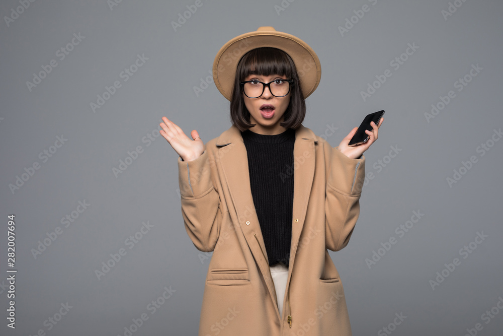 Smiling woman shocked use smartphone in beige coat, round hat on gray background Smiling woman shocked use smartphone in beige coat, round hat on gray background