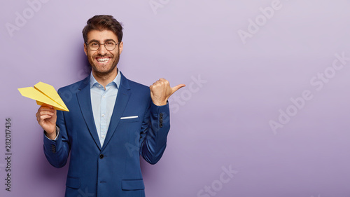 Business success concept. Waist up shot of happy prosperous businessman holds hand made paper airplane, points away with thumb, shows blank space for your promotional content, being in office