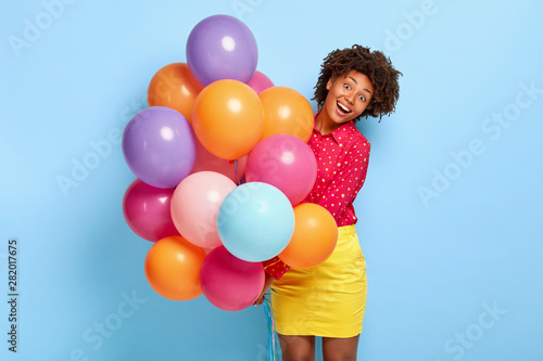 Happiness and celebration concept. Overjoyed dark skinned female greets friends with special occasion  holds huge bunch of helium balloons  has cheerful moments in her life during birthday party
