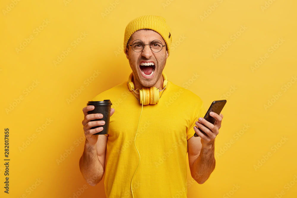 Photo of angry outraged student dressed in yellow outfit, holds smartphone and takeout coffee, screams from negative emotions, reacts on bad news, discontent with something. Monochrome shot. Photo of angry outraged student dressed in yellow outfit, holds smartphone and takeout coffee, screams from negative emotions, reacts on bad news, discontent with something. Monochrome shot.