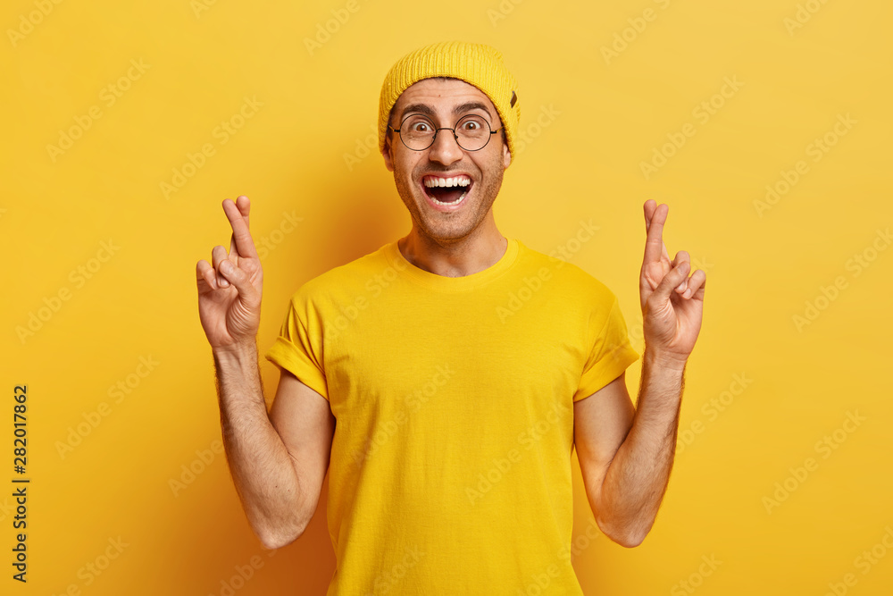 Hopeful cheerful guy crosses fingers for good luck, dreams about something or expects miracle, anticipates good news, wears yellow hat and t shirt makes wish or aims success hopes for excellent result Hopeful cheerful guy crosses fingers for good luck, dreams about something or expects miracle, anticipates good news, wears yellow hat and t shirt makes wish or aims success hopes for excellent result