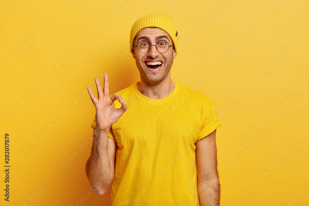 Glad satisfied man shows okay gesture or approval sign, gives positive reply, asserts everything is ok, poses pleased, wears bright vivid clothes, round spectacles, isolated on yellow studio wall. Glad satisfied man shows okay gesture or approval sign, gives positive reply, asserts everything is ok, poses pleased, wears bright vivid clothes, round spectacles, isolated on yellow studio wall.