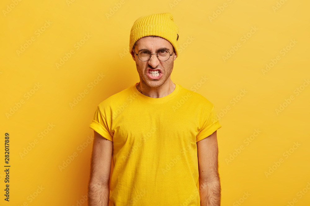 Portrait of irritated man looses temper, clenches teeth from anger, wrinkles nose, looks with hate at camera, says you piss me off, wears transparent glasses, yellow clothes, stands indoor, argues