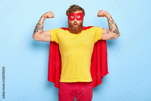 Emotive surprised male hero has noble qualities, demonstrates strength with raised arms, has strong muscles, dressed in superhero costume, isolated on blue, has strength to face dire situation in life