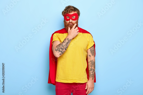 Sad serious superhero man touches thick red beard, wears mask and cloak, looks straightly at camera, thinks about making good things for other people and helping, isolated on blue background.