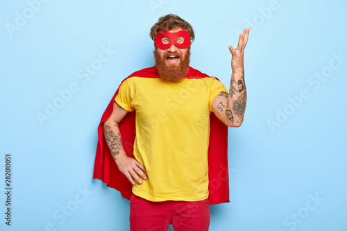 Irritated superhero raises arm and gestures with annoyance, has much work, wears special costume, pretends having supernatural power, outraged to fight with evil, poses indoor. Negative emotions