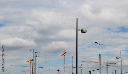 Construction area with lots of tower cranes and street lights with cloudy sly on the background