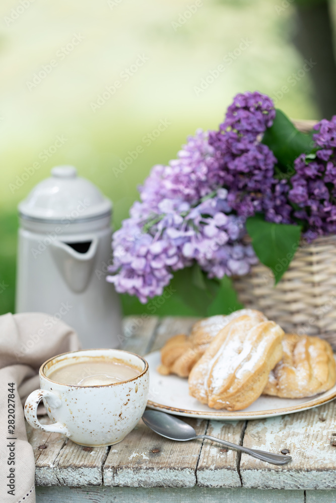 Breakfast in the garden: eclairs, cup of coffee, coffee pot, lilac flowers in a basket.