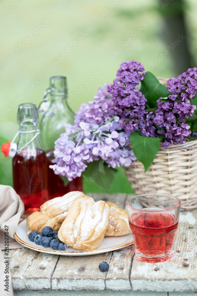 Breakfast in the garden: eclairs, cup of coffee, coffee pot, lilac flowers in a basket.
