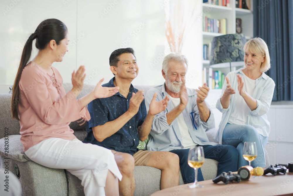 Group of positive seniors European and Asia are singing and drinking wine which sitting on sofa in living room at retirement home. Retired people have smiling and felling happy. Group of positive seniors European and Asia are singing and drinking wine which sitting on sofa in living room at retirement home. Retired people have smiling and felling happy.