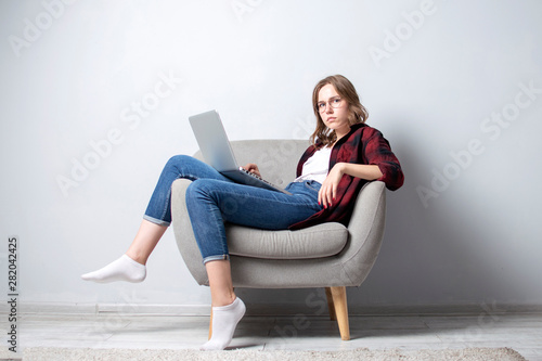 young girl with a laptop sitting on a soft comfortable chair, and relax, a woman using a computer against a white blank wall, she freelancing and printing text, copy space