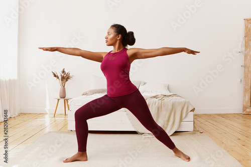 Side view of stylish athletic sporty African girl in leggings and top standing barefooted on floor in warrior 2 pose, doing yoga, strengthening legs, training endurance to get strong healthy body