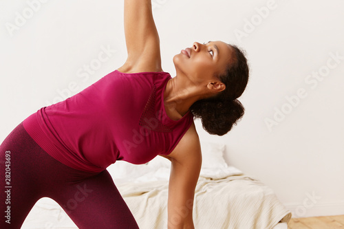 Close up picture of concentrated energetic fit young Afro American woman in stretch top doing warrior 2 yoga stance or virabhadrasana II while exercising at home in the morning. Fitness and activity