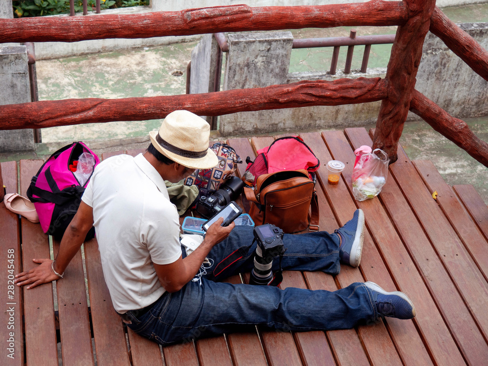 Photographer is sitting on the wood bridge. Cameraman is checking photos in his mobile phone. Man is traveler. All around of him is bags and camera with lens. Male is wearing white shirt, jean, hat. Photographer is sitting on the wood bridge. Cameraman is checking photos in his mobile phone. Man is traveler. All around of him is bags and camera with lens. Male is wearing white shirt, jean, hat.