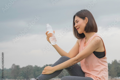 Beautiful asian woman drinking water after work out exercising at summer green park.