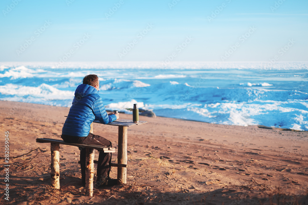 A man sits on a bench and looks into the horizon. Beach in the winter. Thermos on the table
