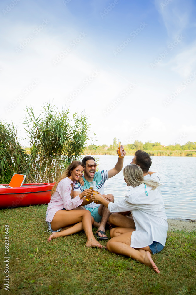 Group of friends with cider bottles sitting by the boat near the beautiful lake and having fun