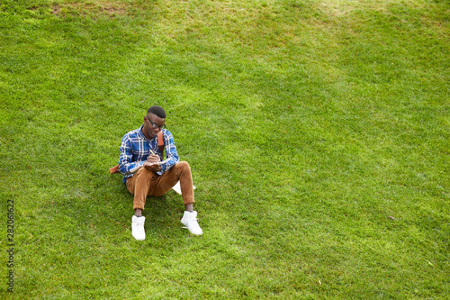 Wide angle portrait of young African-American man sitting on green grass in campus and doing homework outdoors, copy space