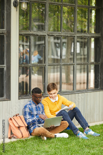 Full length portrait of two cheerful international students studying together outdoors while sitting on green grass in college campus, copy space