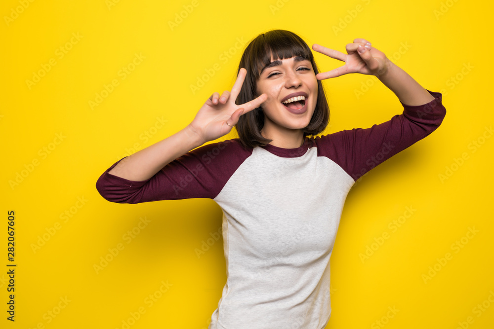 Happy pretty girl posing at camera showing peace sign isolated on yellow background Happy pretty girl posing at camera showing peace sign isolated on yellow background