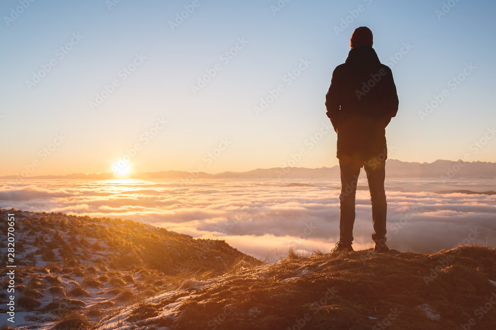 View From the back. A lonely standing man high in the mountains looks at the setting sun and the sunset horizon with a valley filled with clouds. The concept of tourism travel and male loneliness - obrazy, fototapety, plakaty 