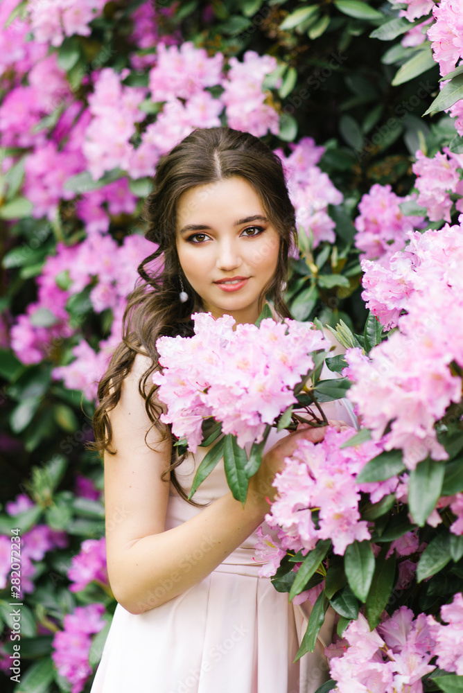 Beautiful portrait of a lovely girl who stands surrounded by lilac pink flowers blooming rhododendron in the spring in the garden Beautiful portrait of a lovely girl who stands surrounded by lilac pink flowers blooming rhododendron in the spring in the garden